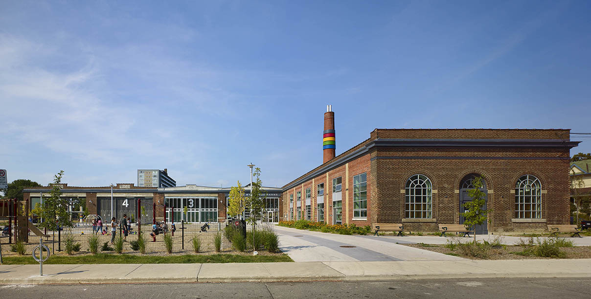 Kids play on a playground in front of five side-by-side red-brick warehouses